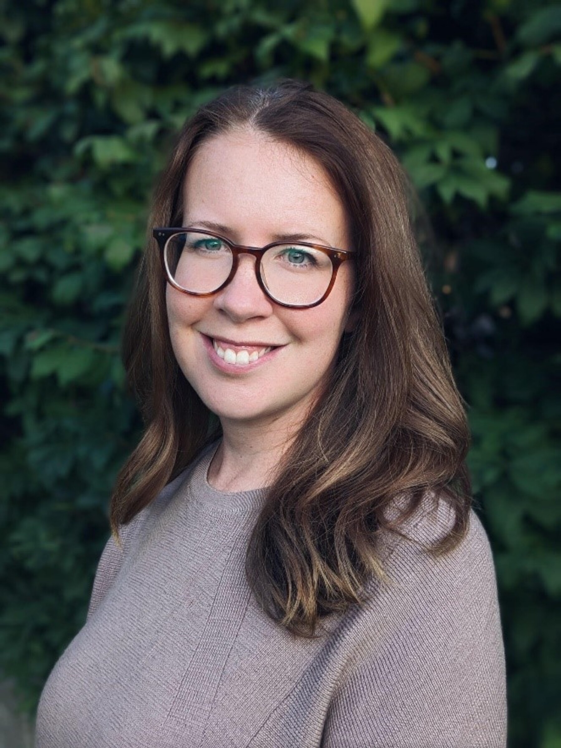 Photo of Dr. Kathleen Fraser from the shoulders up. She is outdoors with greenery in the background. She has wavy dark brown hair past her shoulders. She has brown-rimmed glasses. She is smiling at the camera.