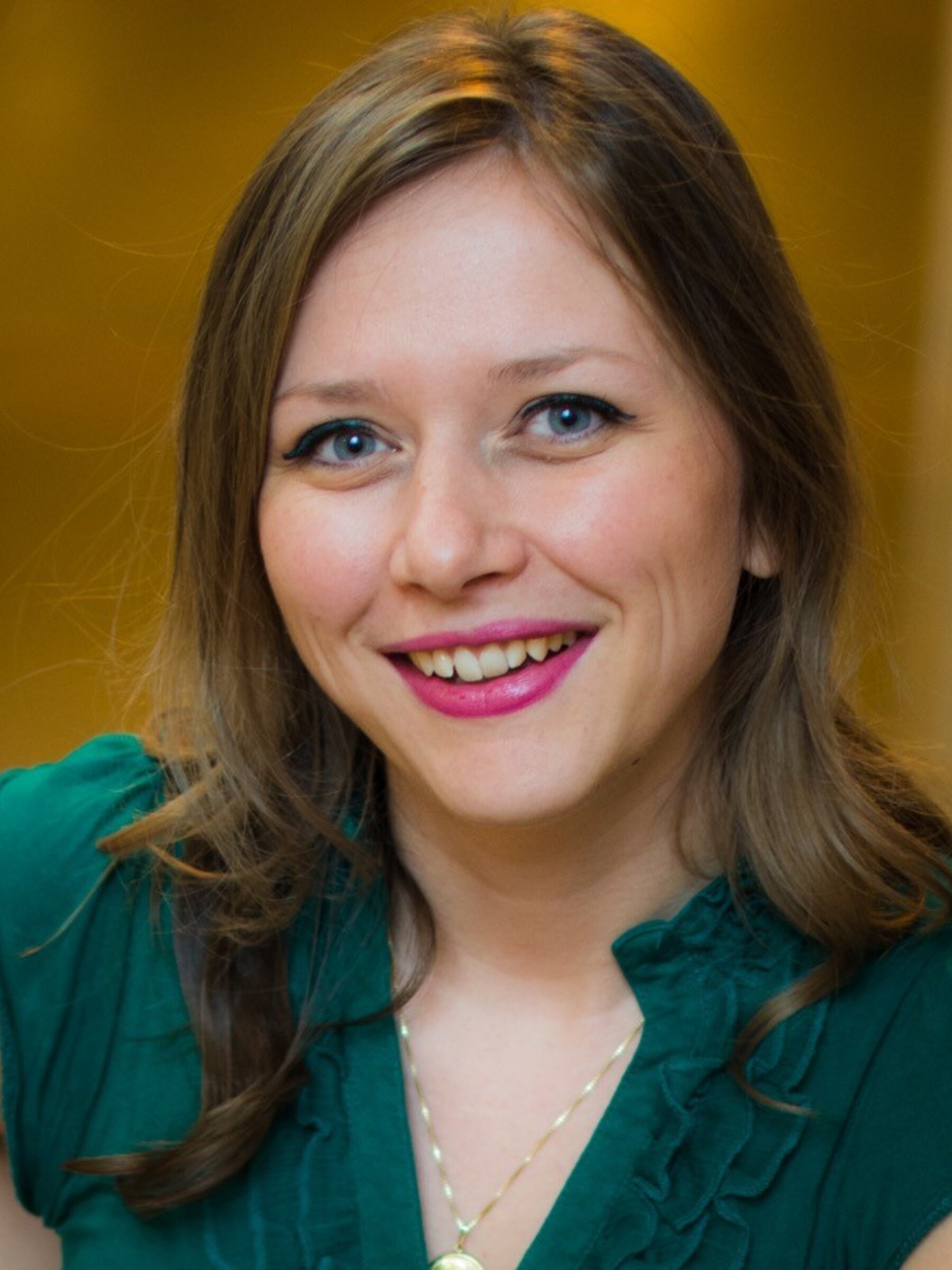 Photo of Dr. Tijana Simic from the shoulders up. She is wearing a forest green blouse. She is smiling at the camera. She has wavy brown hair past her shoulders and blue eyes. She has pink lipstick on.